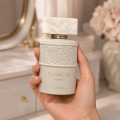 A hand holding a white perfume bottle labeled 'PANACHE Angel Dust' by the brand 'Khadlaj'. The bottle features an embossed floral pattern on the upper half and a smooth white cap, set against a softly blurred vanity background.