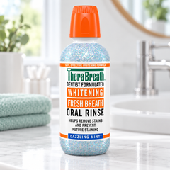 A bottle of TheraBreath Fresh Breath Whitening Oral Rinse in Dazzling Mint, featuring a white label and bright orange cap, set on a marble bathroom counter with a sink and faucet in the background.
