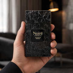 A hand holding a textured black rectangular perfume bottle labeled 'Noiré ABSOLU ARABIYAT PRESTIGE' against a blurred indoor background.