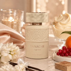 A frosted cream perfume bottle labeled 'PANACHE Angel Dust' by Khadlaj, styled on a marble surface with white flowers, vanilla beans, a lit candle, whipped cream, and red berries.